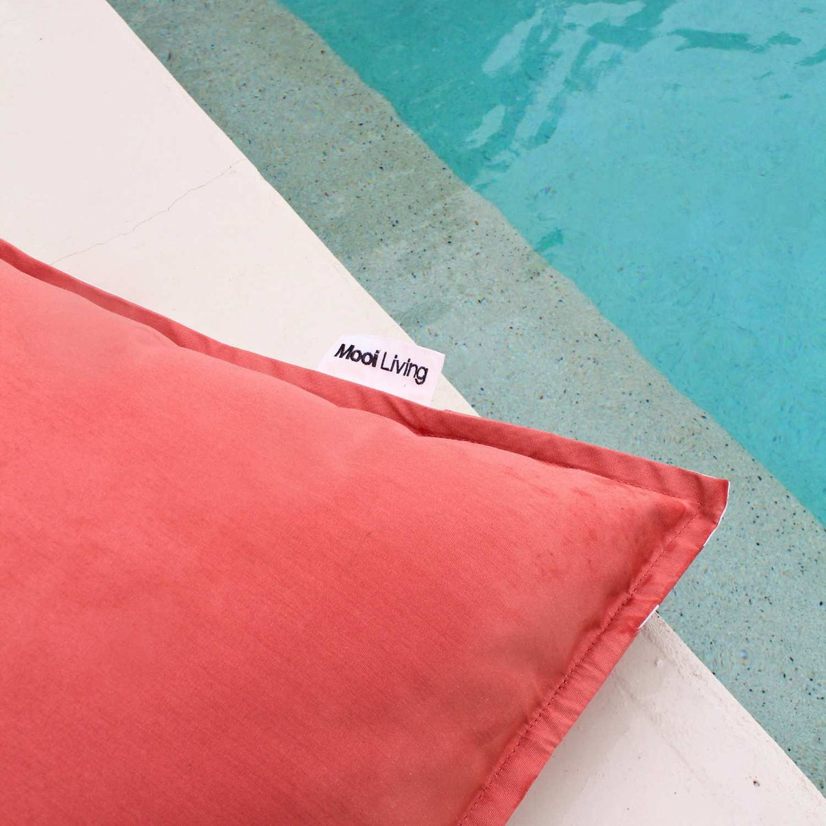 Close-up of the plain blush fabric, stitched edge and Mooi Living label on a reversible triangle outdoor bean bag beside a pool, MOREVSTRIBB-BLUS