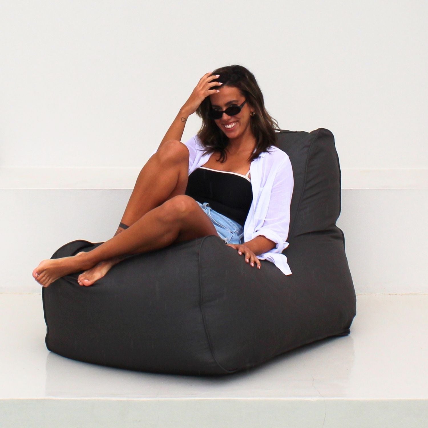 A woman wearing sunglasses sits on a Oyster Black Bora Outdoor bean Bag Chair in a minimalist white room, MOBORABB-OYSBLA
