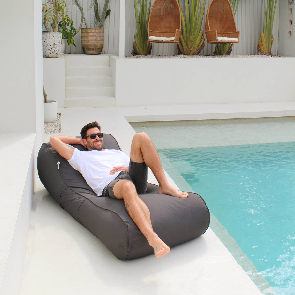 Angled view of a man reclining on a Mooi Living Oyster Black Isla outdoor bean bag sunlounger on a white poolside terrace, MOISLABB- OYSB
