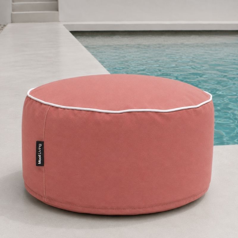 A Blush Outdoor Ottoman with white piping and a small black 'Mool Living' tag, positioned next to a swimming pool on a light concrete patio.
