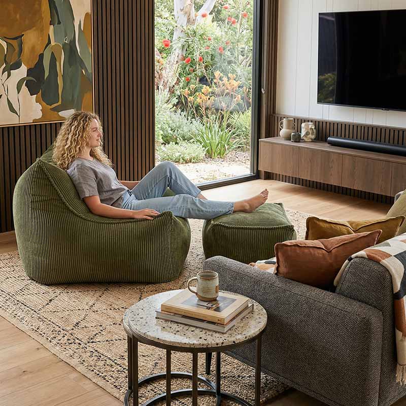 Lifestyle image of the Mooi Cord Bean Bag Chair indoor bean bag by Mooi Living in Olive Green with a woman relaxing on the bean bag chair in a modern living room.