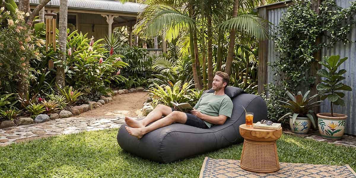 Australian Tropical backyard - Lush green garden with grass, trees and soft landscaping, Oyster Black Isla sunlounger outdoor bean bag by Mooi Living. Bean bag placed on the lawn in dappled sunlight.