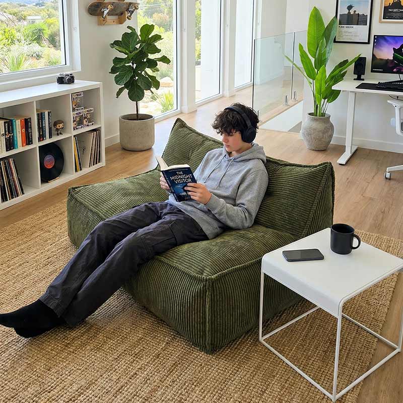 Lifestyle image of the Mooi Double Cord indoor bean bag by Mooi Living in Olive Green with a teen sitting on it while listening to music and reading a book in a modern living space.