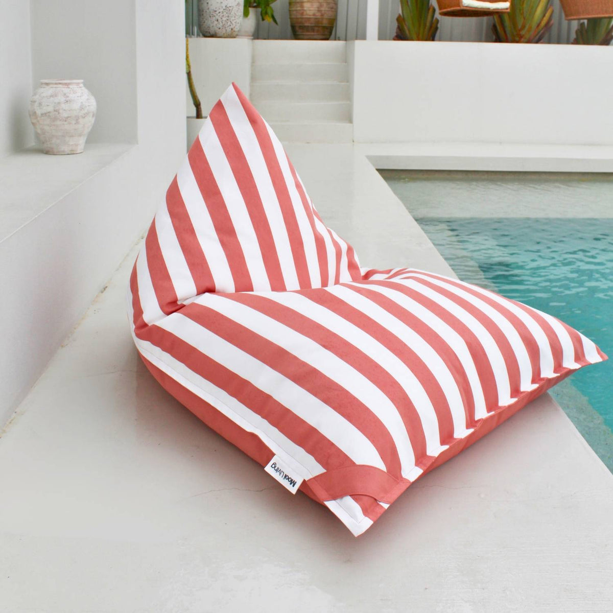 Mooi Living Blush reversible triangle outdoor bean bag beside a pool, showing its blush and white striped side, MOREVSTRIBB-BLUS