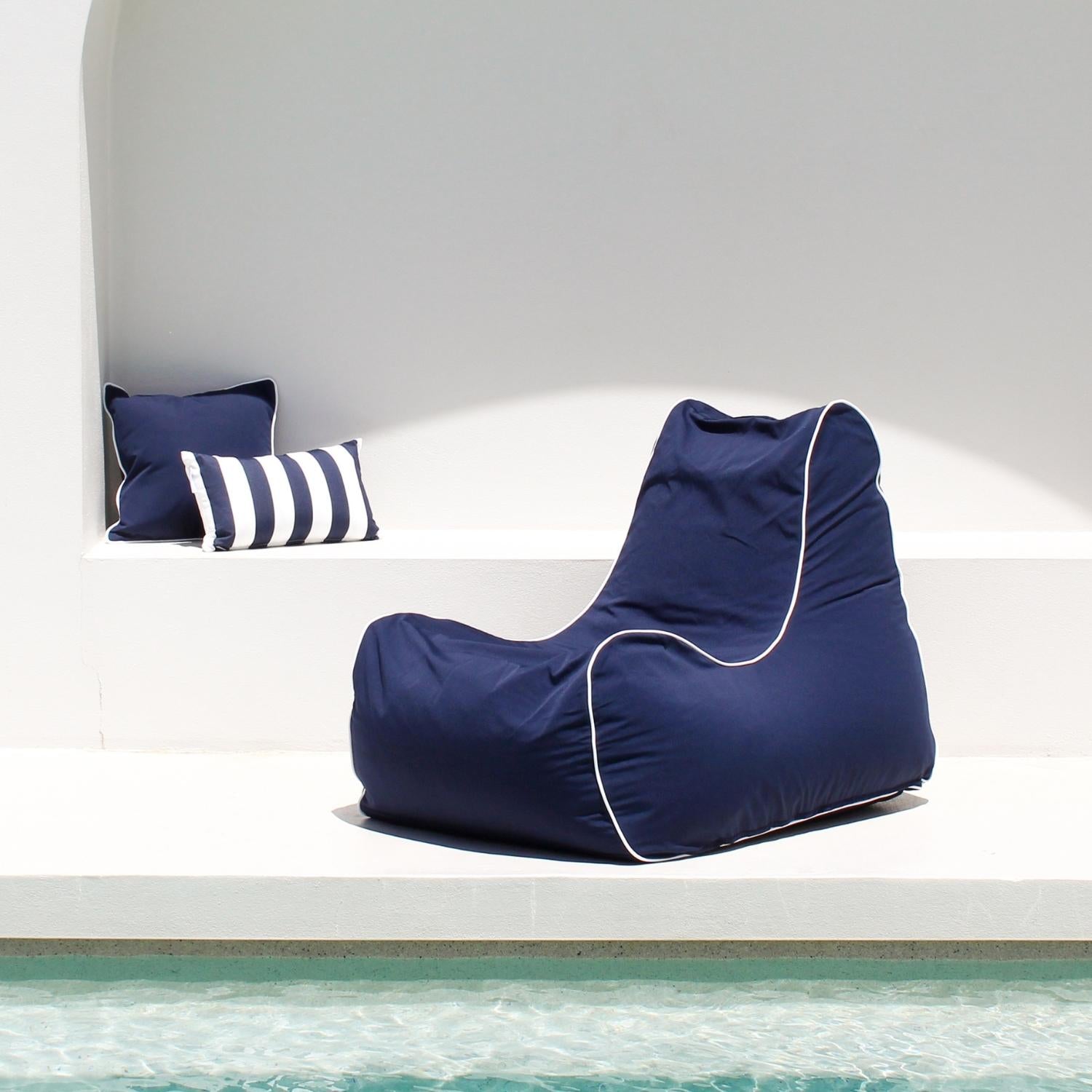 A navy Bora Bora bean bag chair with white piping sits on a white patio ledge next to a swimming pool, with navy and navy stripes pillows resting in a recessed shelf area behind it.