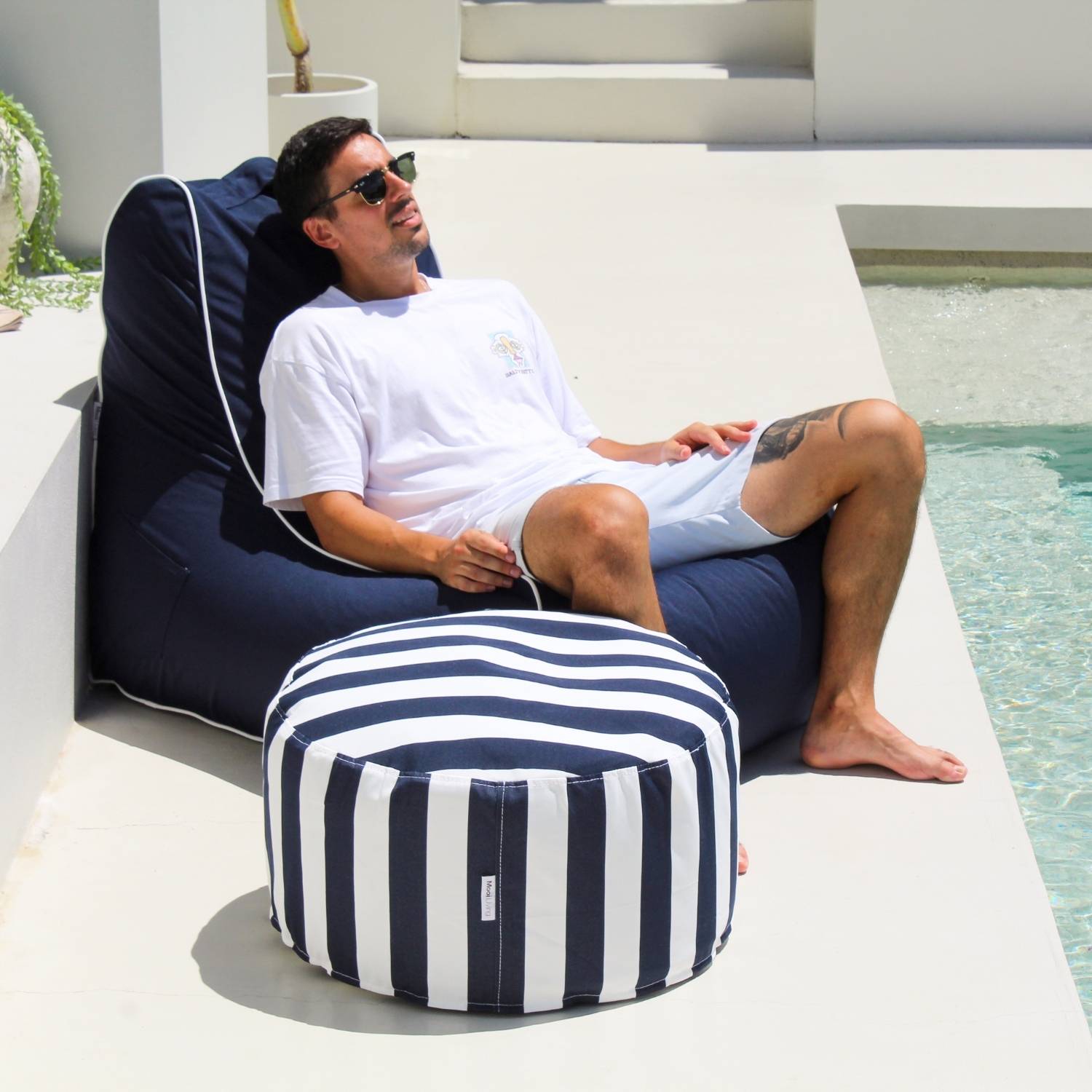 A man relaxes in a navy Bora Bora outdoor bean bag chair next to a swimming pool, with a matching navy and white striped outdoor ottoman in the foreground.