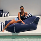 A woman wearing sunglasses reclines on a large navy Chill UV  bean bag chair with white piping by Mooi Living, MOCHUVBB-NAVY.