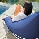 Rear close-up of a woman reclining on the plain navy side of a reversible triangle outdoor bean bag on a poolside deck, MOREVSTRIBB-NAVY