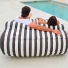 Rear view of a couple sharing an Oyster Black Nevaeh double outdoor bean bag sunbed facing a pool, showing its striped back and bolster cushion, MOSTNEVBB-OYSBLA