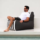 A man wearing sunglasses, a white t-shirt, and dark shorts reclines on a Mooi Living Oyster Black Stripe Bora Bora bean bag chair, positioned next to a swimming pool against a clean white wall,MOBORABB-STRI-OYSBLA