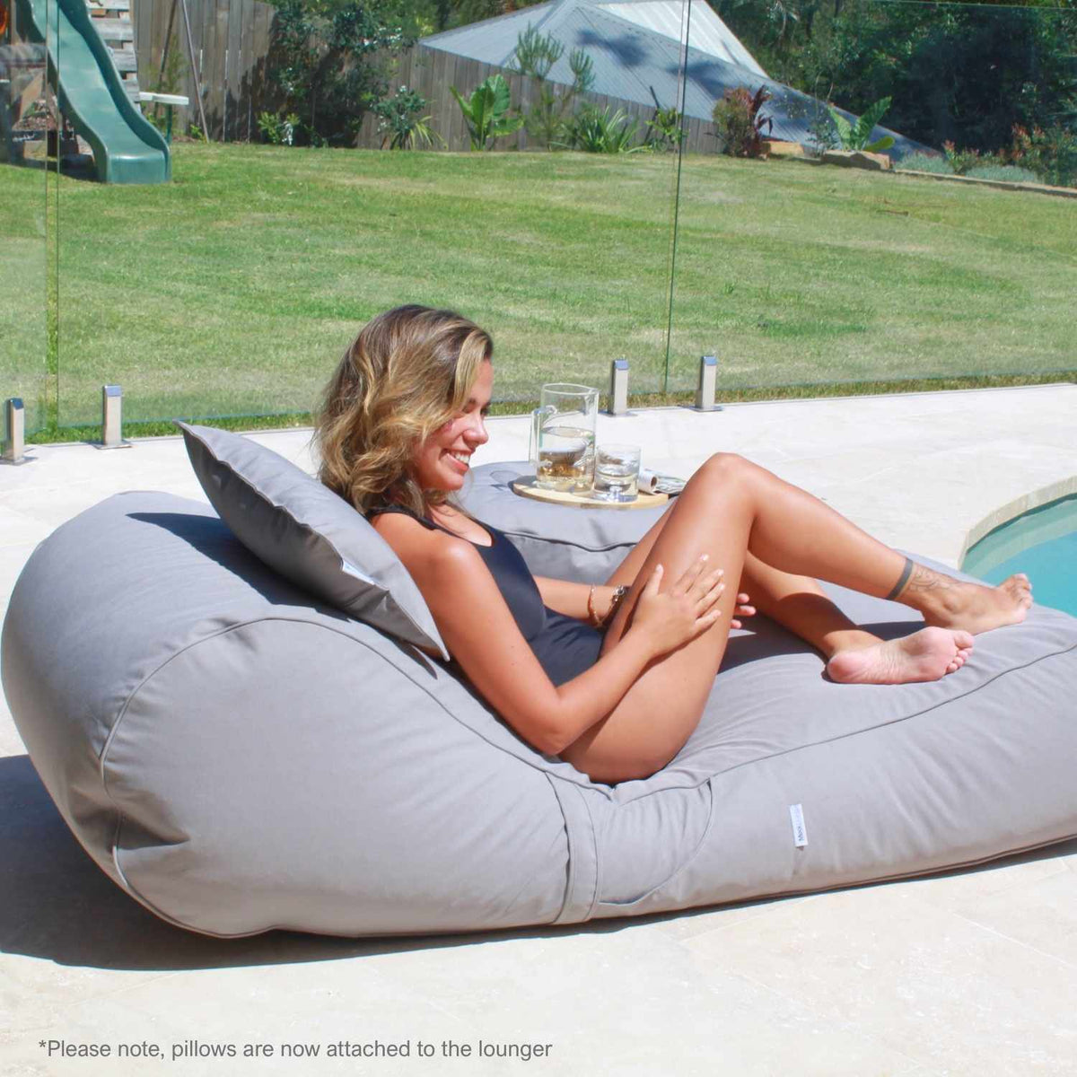 Side view of a woman lounging on a Mooi Living Pumice Stone Isla outdoor bean bag sunlounger beside a pool, showing its curved backrest and pillow, MOISLABB-PUMSTO