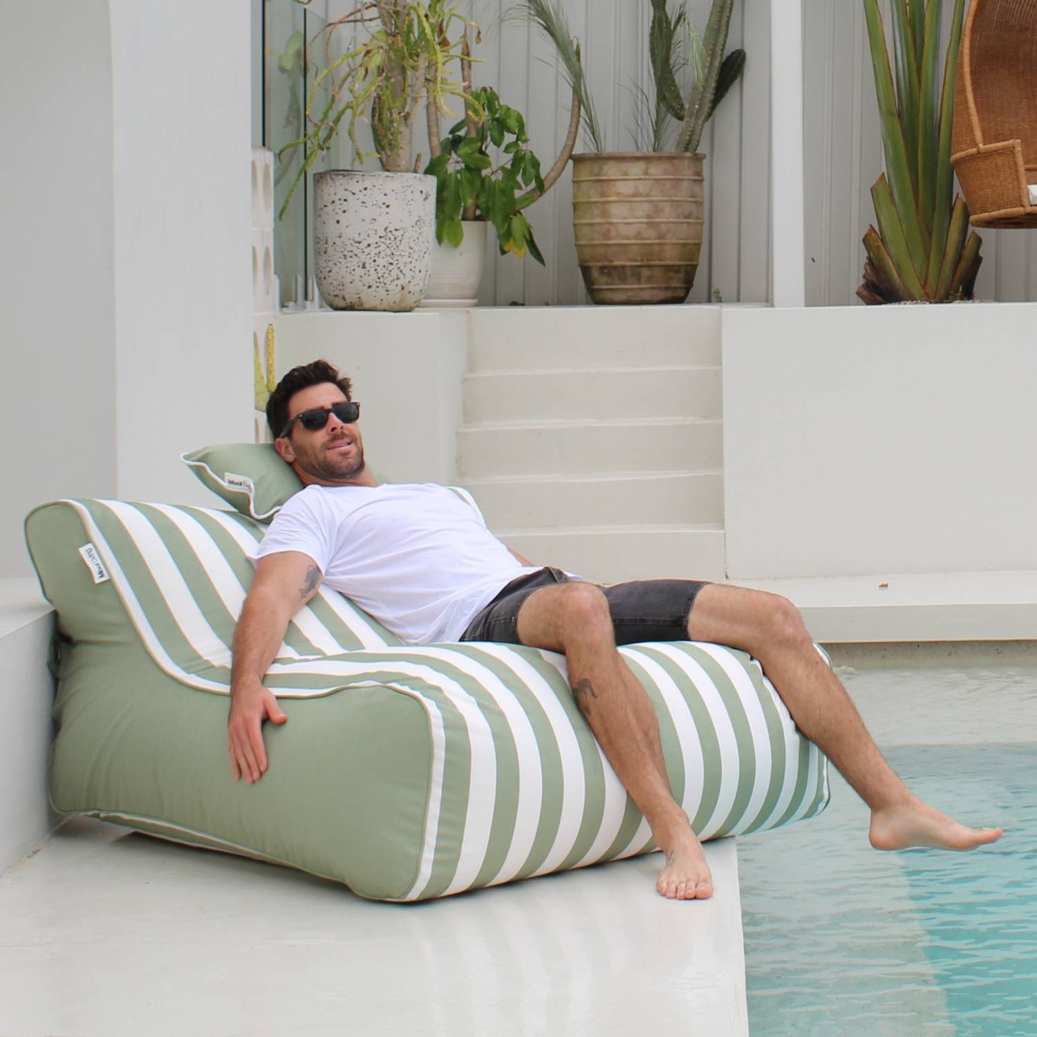 A man wearing sunglasses relaxes on a sage green striped Chill UV outdoor lounger by Mooi Living, next to a swimming pool, MOSTRCHILL-BB-GRESTR.