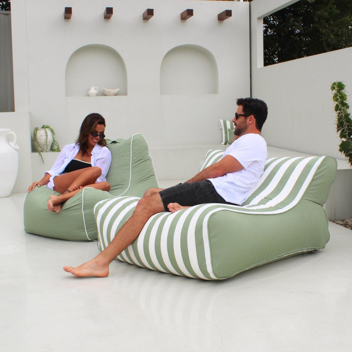 A man and a woman relax on a Bora Bora outdoor bean bag lounge chairs with sage green and white stripes in a modern white patio setting, MOSTRCHILL-BB-GRESTR