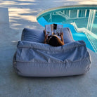 Rear view of a woman reading on a Mooi Living Pumice Stone Nevaeh double outdoor bean bag sunbed, showing its backrest and bolster cushion beside a pool, MOSTNEVBB-PUMSTO