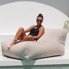 A woman wearing a black swimsuit and sunglasses sits on a large, taupe Chill UV outdoor lounge bean bag chair with white piping, positioned next to a white pool deck under a bright sun.