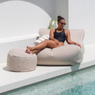A woman in a black swimsuit and sunglasses relaxes on a taupe Chill UV outdoor lounger next to a pool, with a matching round ottoman and a striped pillow in a modern white patio setting.