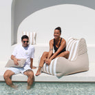 A man and a woman relax by a poolside on oversized beige and white striped bean bag loungers, with a white modern architectural background, MOBORABB-STRI-TAU