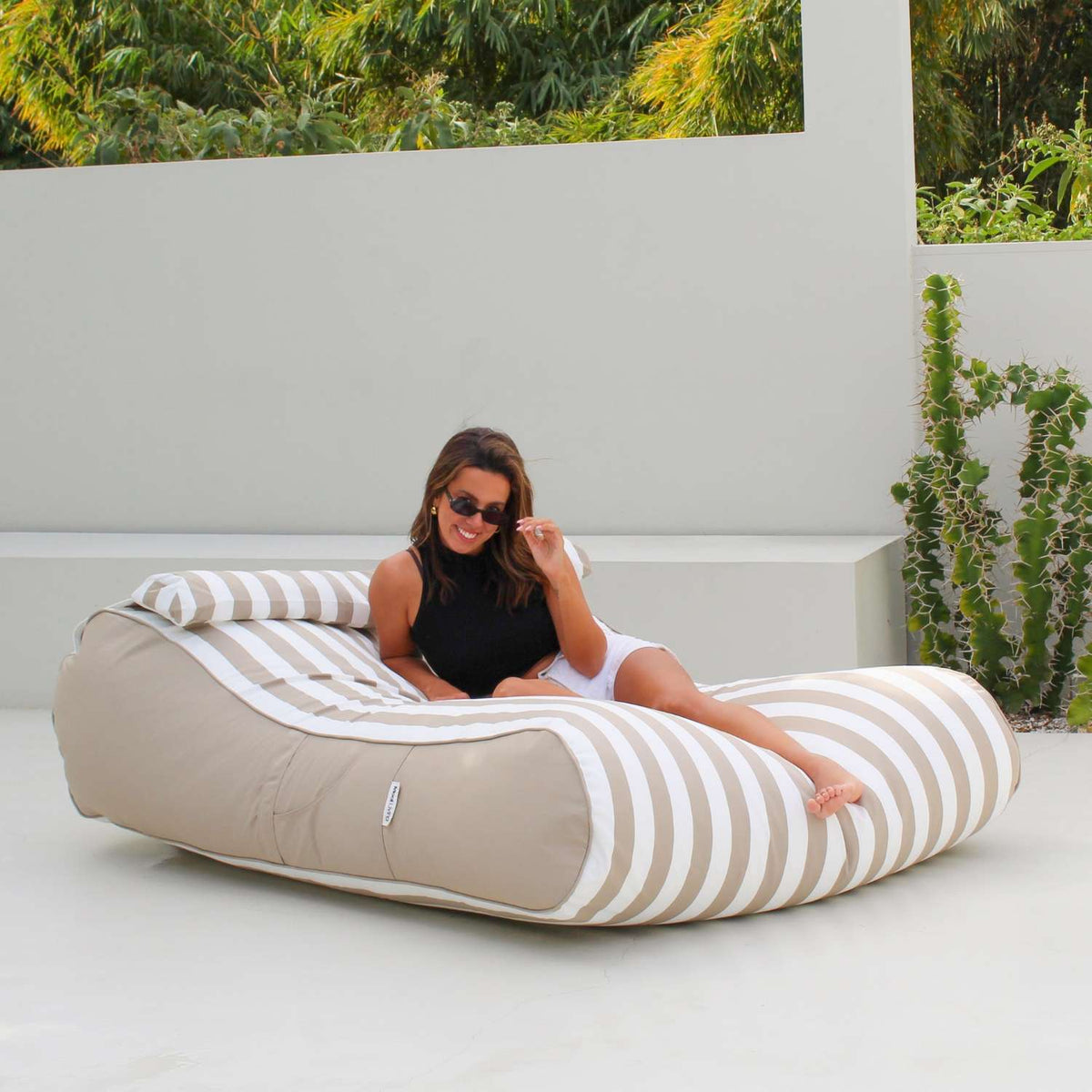 Woman sitting on a Mooi Living Taupe Stripe Nevaeh double outdoor bean bag sunbed on a white patio beside cacti, MOSTNEVBB-STRI-TAU