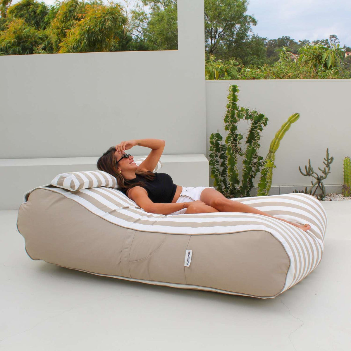 Side view of a woman reclining on a Mooi Living Taupe Stripe Nevaeh double outdoor bean bag sunbed with a matching bolster cushion, MOSTNEVBB-STRI-TAU