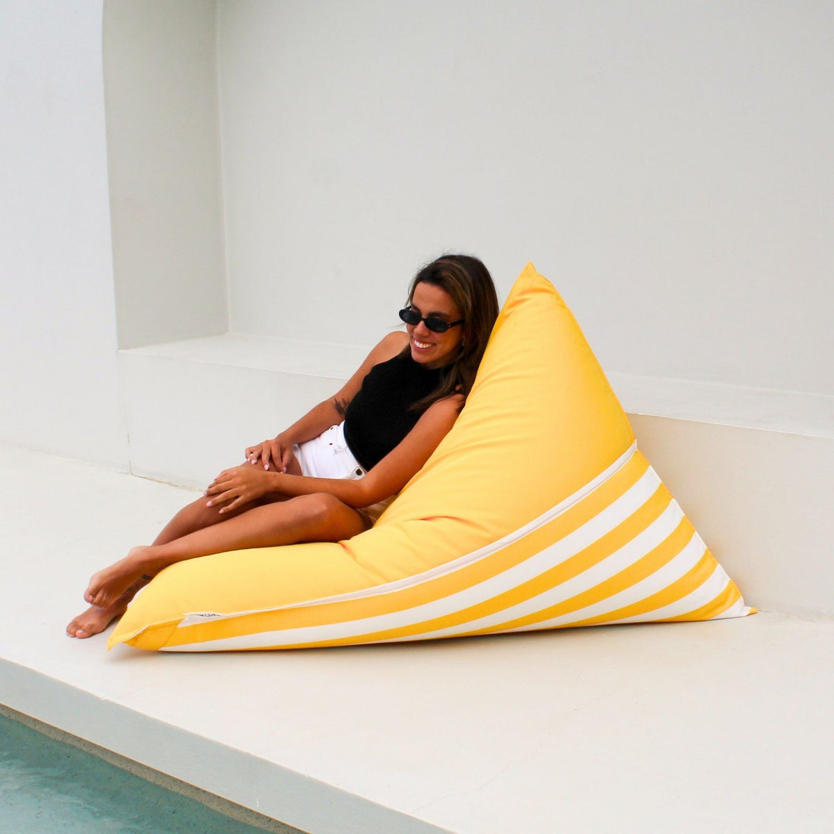 Woman relaxing on the yellow and white striped side of a Mooi Living reversible triangle outdoor bean bag beside a pool, MOREVSTRIBB-YEL