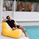 A man wearing sunglasses and a black t-shirt relaxes on a bright yellow Bora bean bag chair with white stripes next to a swimming pool in a modern outdoor patio setting, MOBORABB-STRI-YEL