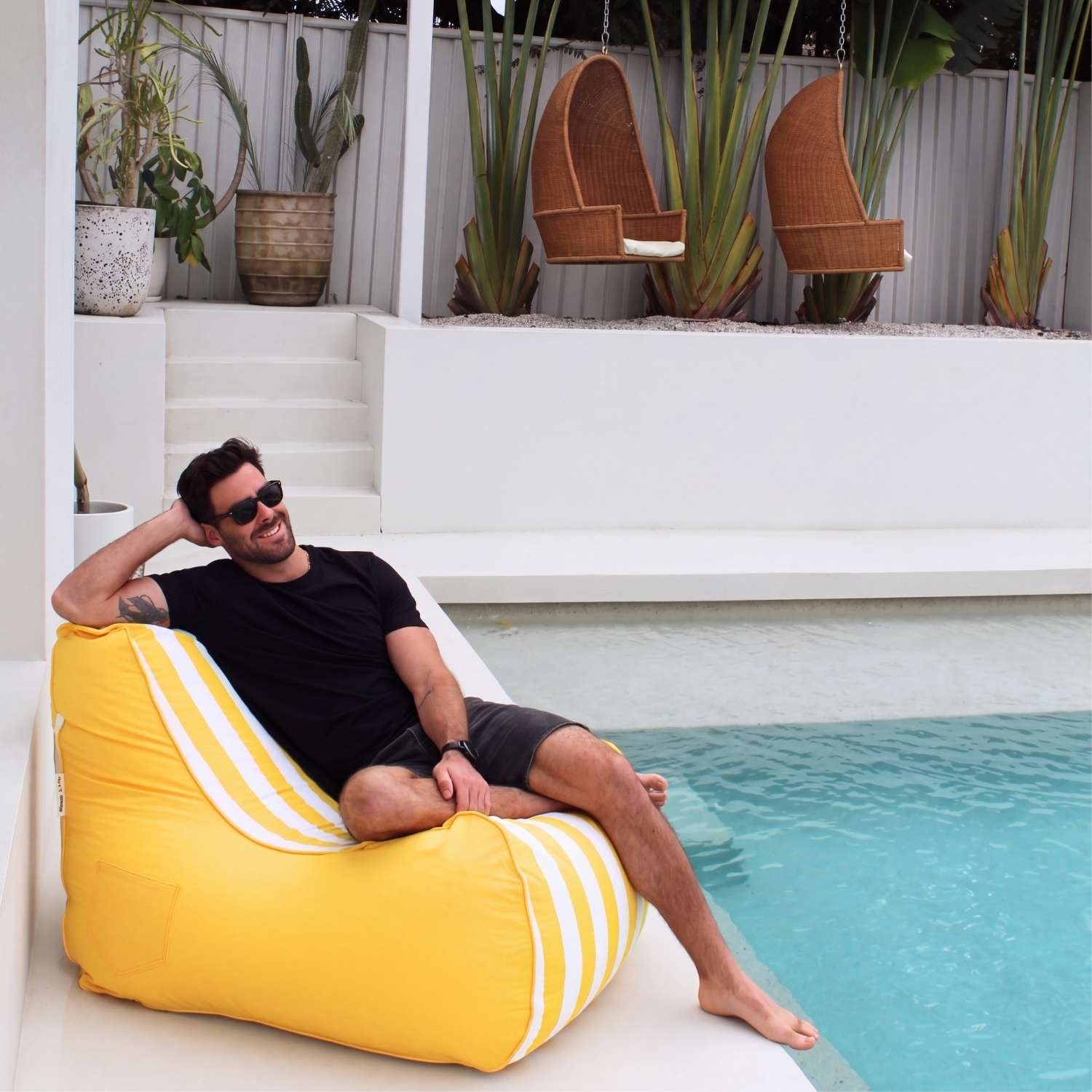 A man wearing sunglasses and a black t-shirt relaxes on a bright yellow Bora bean bag chair with white stripes next to a swimming pool in a modern outdoor patio setting, MOBORABB-STRI-YEL