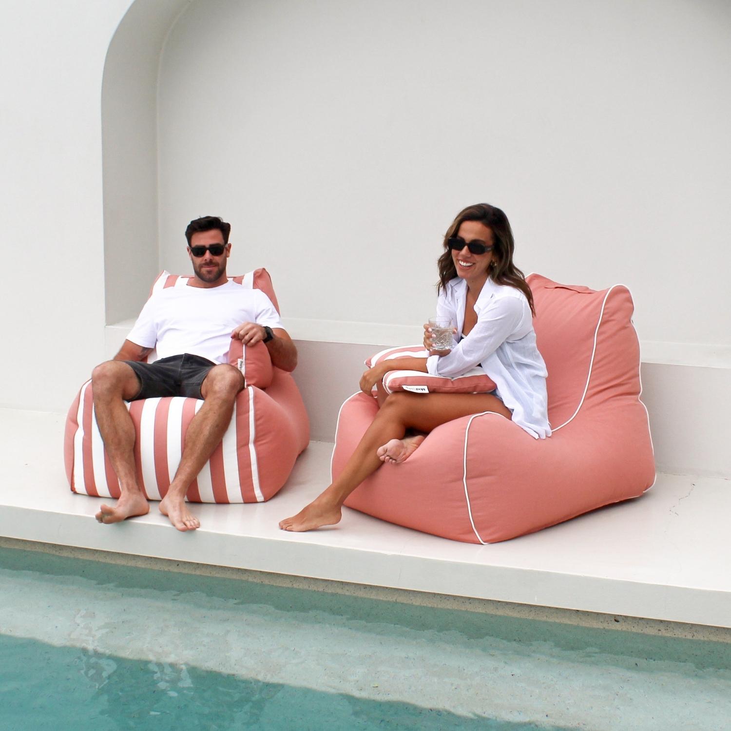 A man and a woman relax in Blush Bora Bora bean bag chairs by a pool. The man sits in a blush and white striped bean bag chair, while the woman sits in a solid blush bean bag chair with white piping,MOBORABB-BLUSH