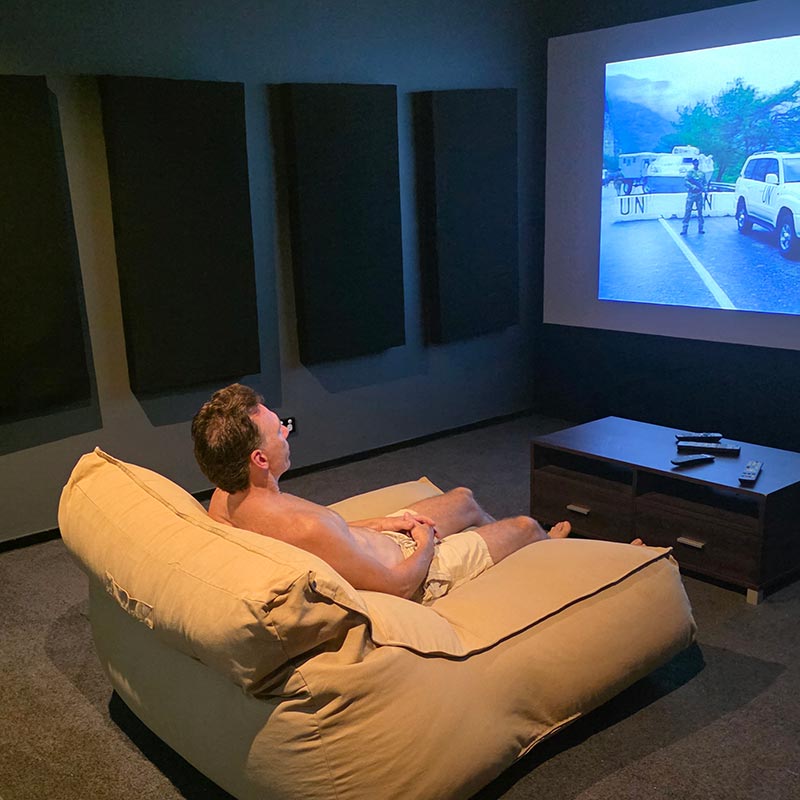 Customer pic of the Double Boss Lounger in Tan. A man is sitting on the Double Boss watching TV in a dark room with a projector screen displaying a scene.