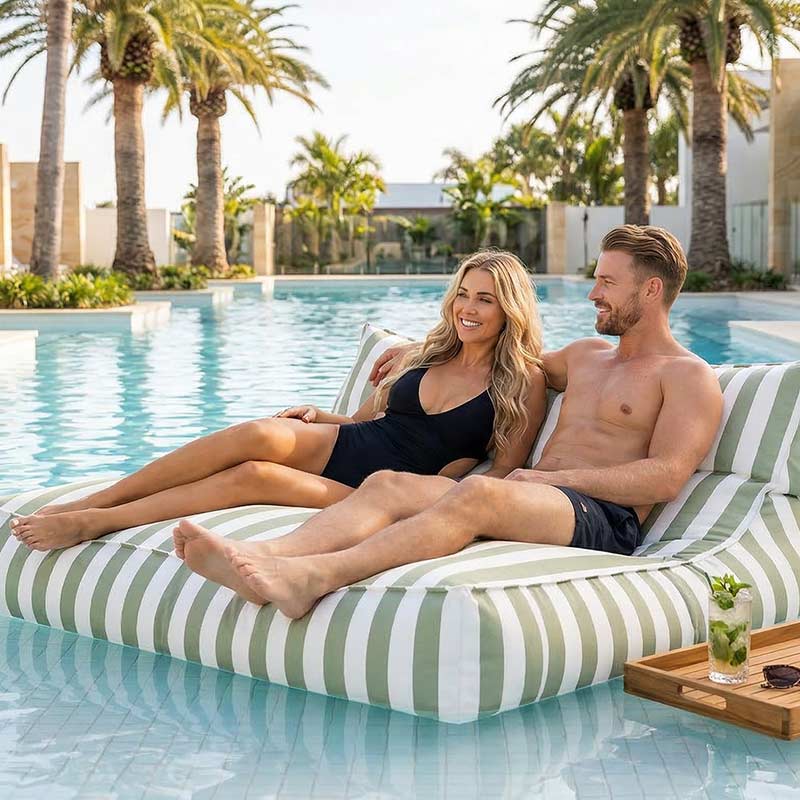 Lifestyle Pic - Luxury resort-style pool in Australia with palm trees, white tiles and clear blue water, two adults relaxing together on a green stripe  Lana double seater bean bag outdoor bean bag by Mooi Living floating on the pool, drinks nearby.
