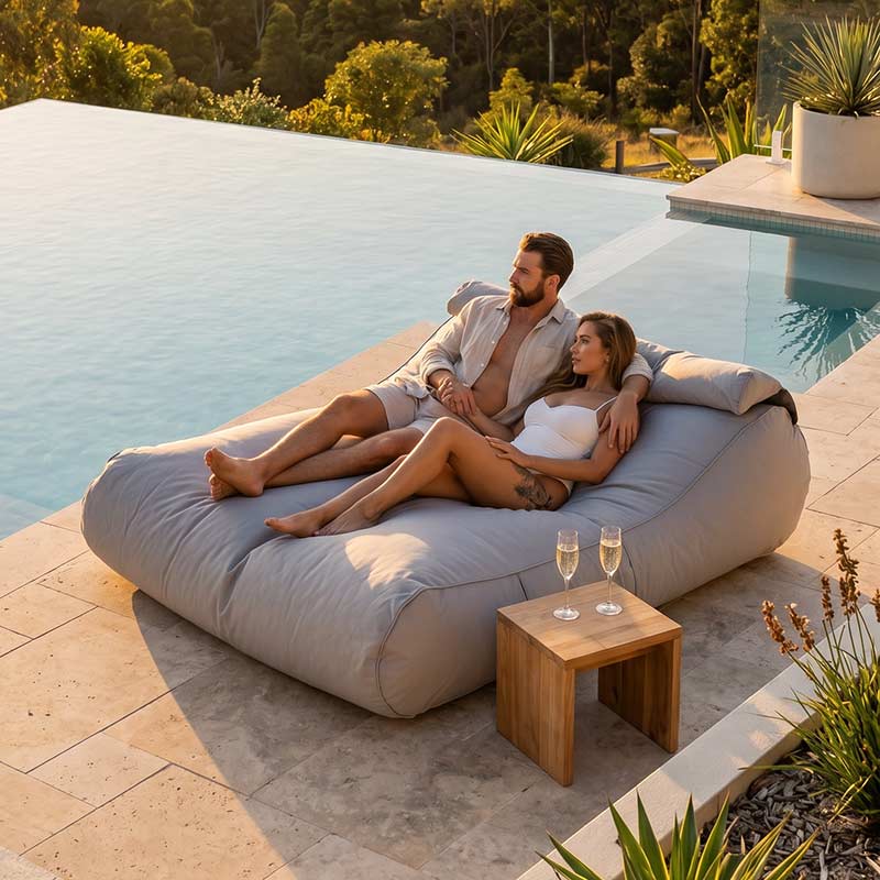 Couple relaxing on a Pumice Stone Nevaeh Sunbed outdoor bean bag by Mooi Living by a pool with a wooden table and glasses.