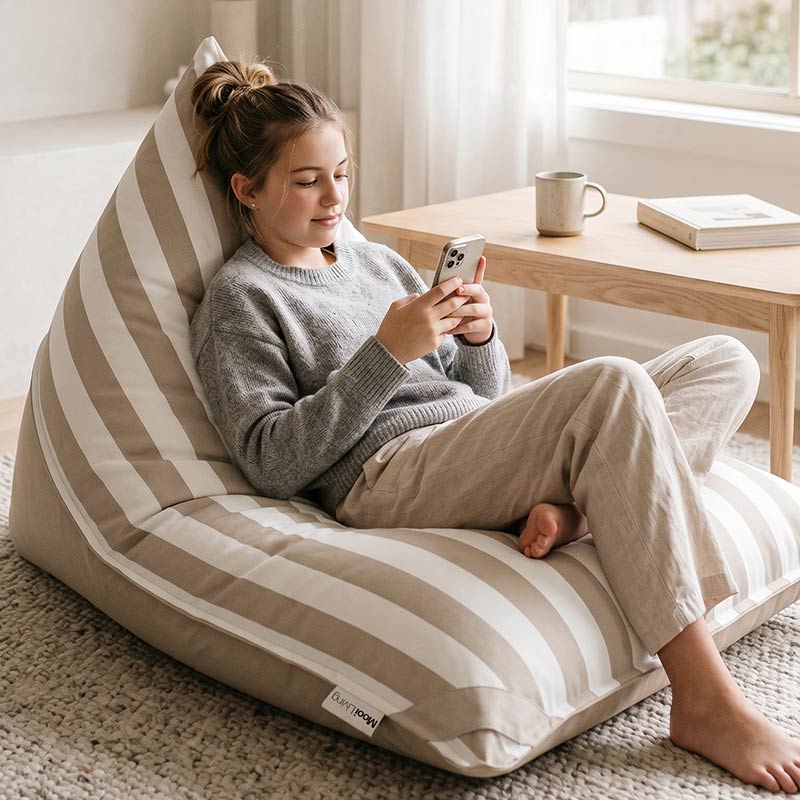 Young woman sitting on a Taupe Reversible Triangle outdoor bean bag by Mooi Living styled in an indoor cozy room while using her phone.