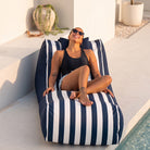 A woman wearing sunglasses and a black swimsuit lounges on a navy blue and white striped Cojelo Suave Sunlounge outdoor bean bag next to a swimming pool.